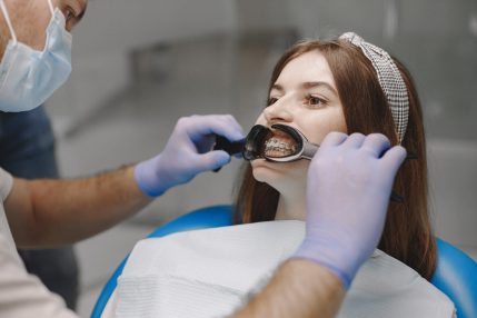 Female patient with braces has dental examination at dentist office. Woman wearing white clothes. Male dentist treats girl.